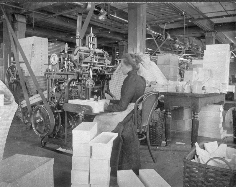 Young woman in long black dress with light-colored half apron seated at envelope-making machine. Long Pulley off overhead shaft very clearly visible. Someone not contemporary with the photograph has suggested this may be Nellie Carmen (could have been Rachael Black's suggestion). — Nellie Carmen envelope maker
